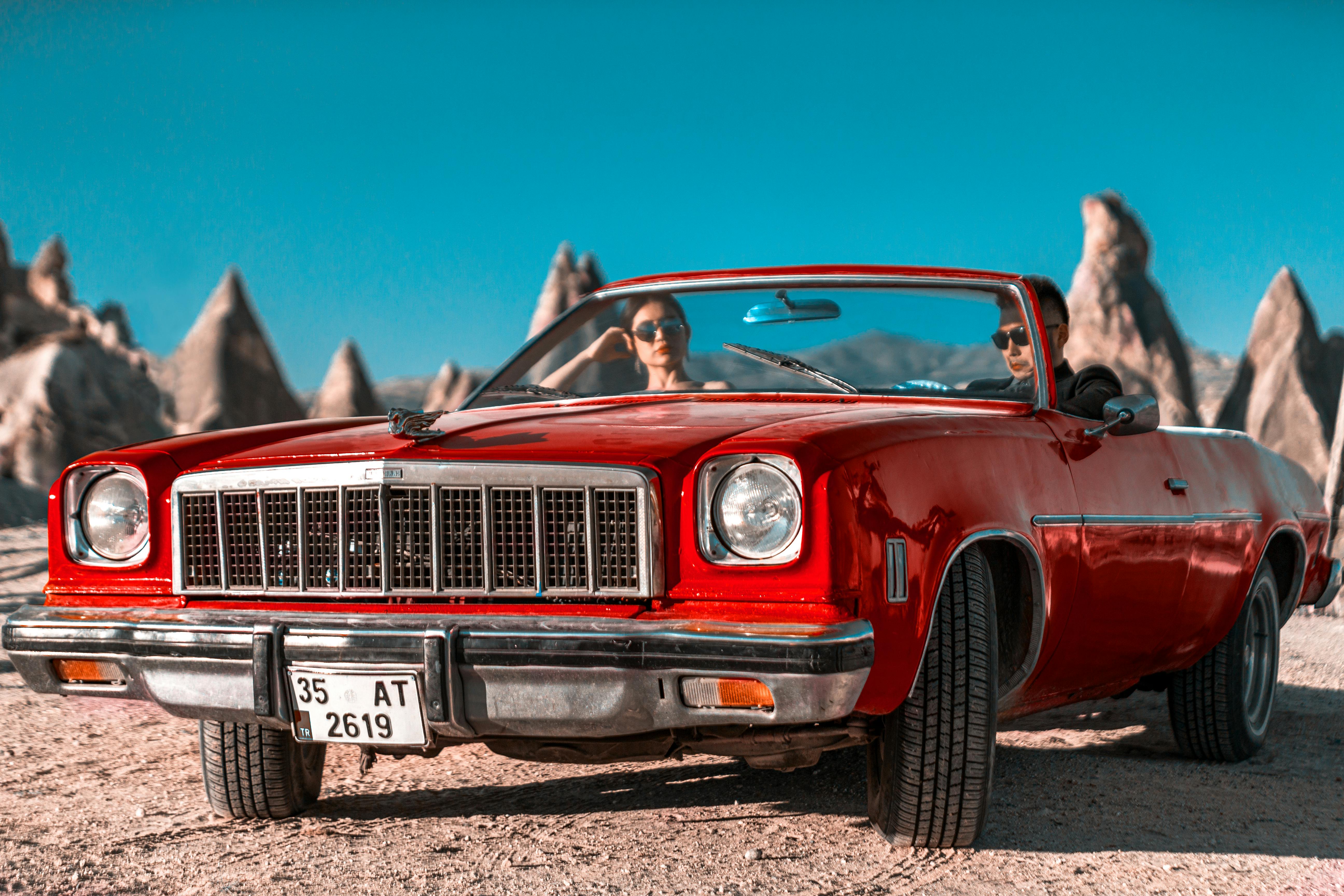 Vintage red convertible car with couple in Cappadocia's unique rock formations under a clear blue sky.