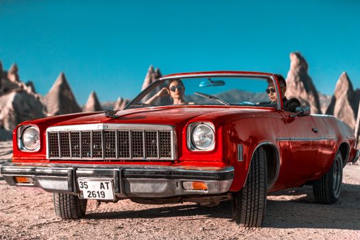 Vintage red convertible car with couple in Cappadocia's unique rock formations under a clear blue sky.