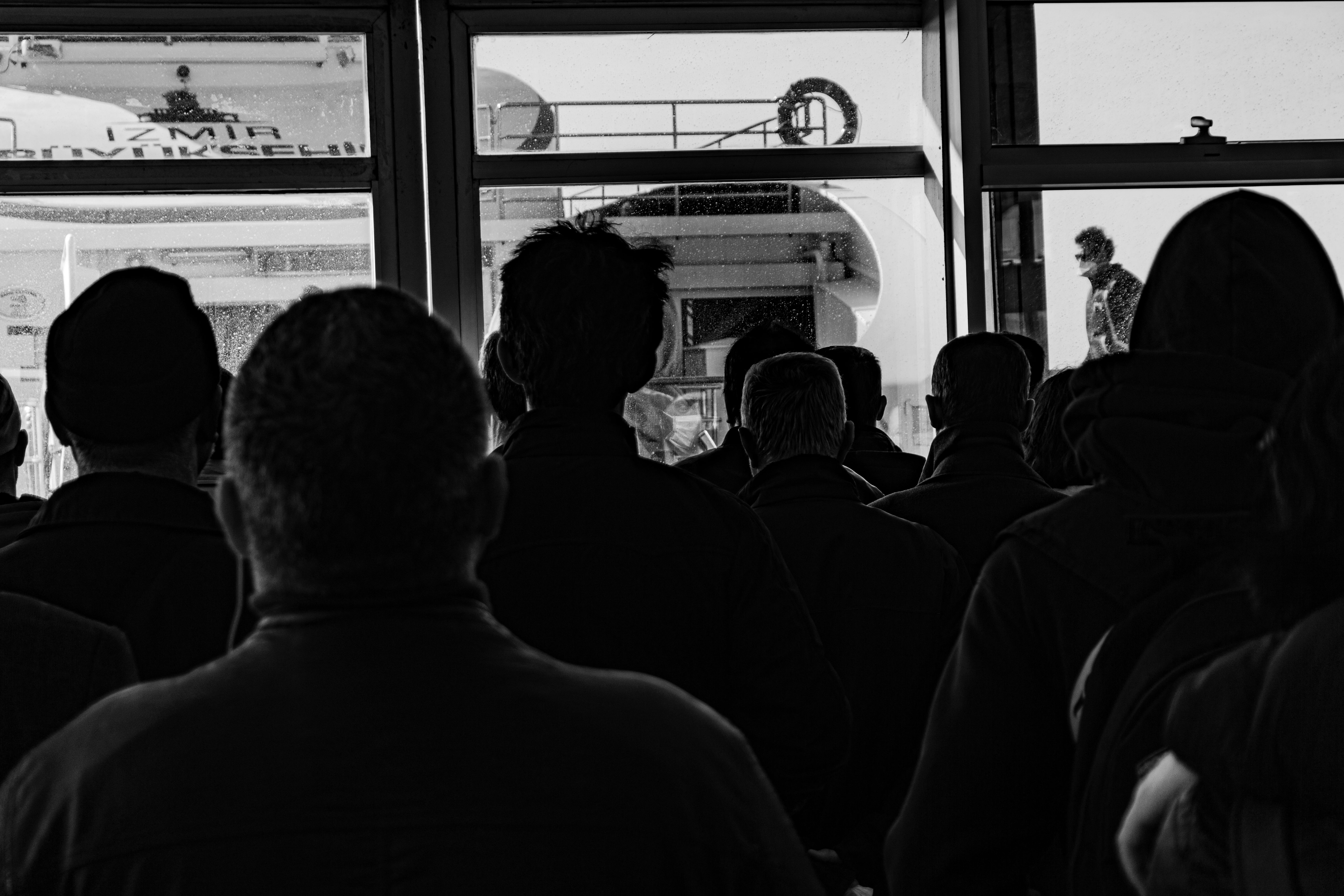 Black and white image of silhouettes indoors, facing a ferry through a glass door.