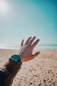 A hand with a watch reaches out towards the ocean on a sunny beach day, capturing summer vibes.