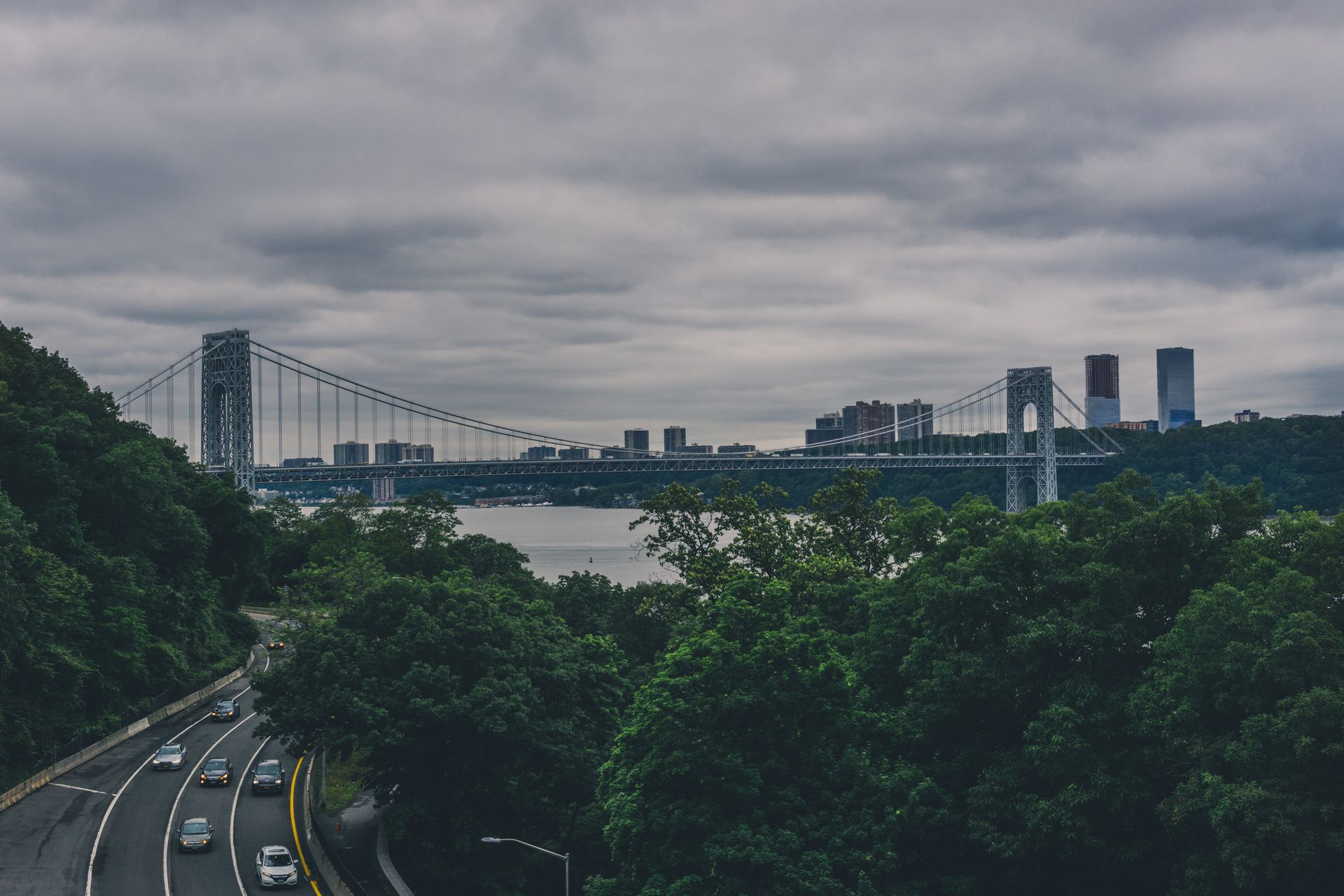 Suspension bridge over urban river with cars and city skyline under a cloudy sky.
