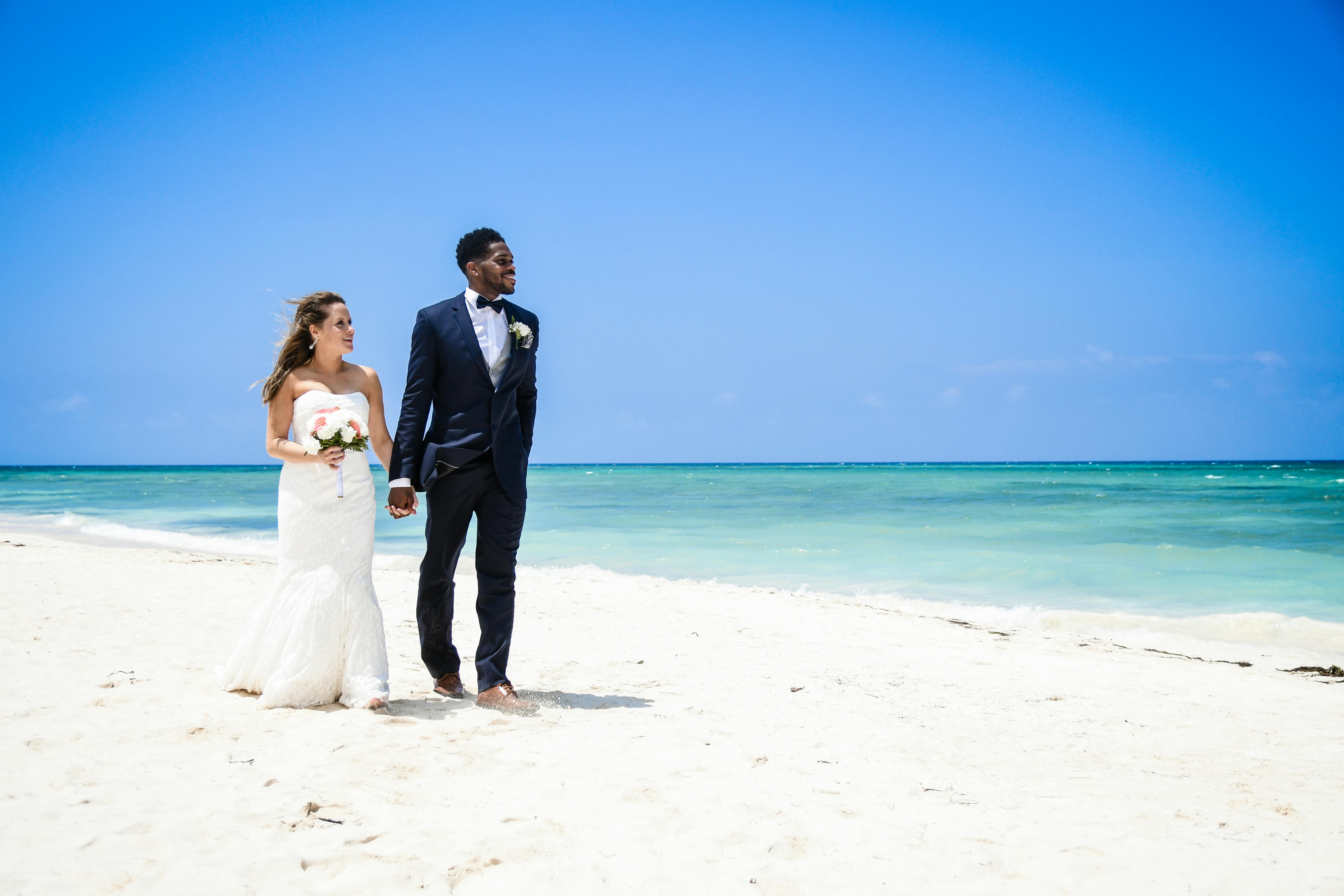 Interracial couple walking hand in hand on a beautiful beach, celebrating their wedding day.
