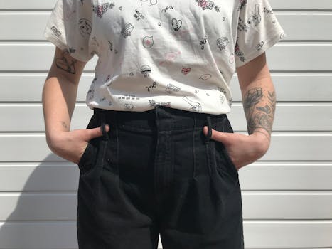 Close-up of a person wearing a printed shirt and black pants with tattoos, standing against a white background.