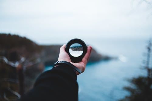 Crop unrecognizable traveler holding modern loupe in hand and admiring picturesque ocean and rocky formations