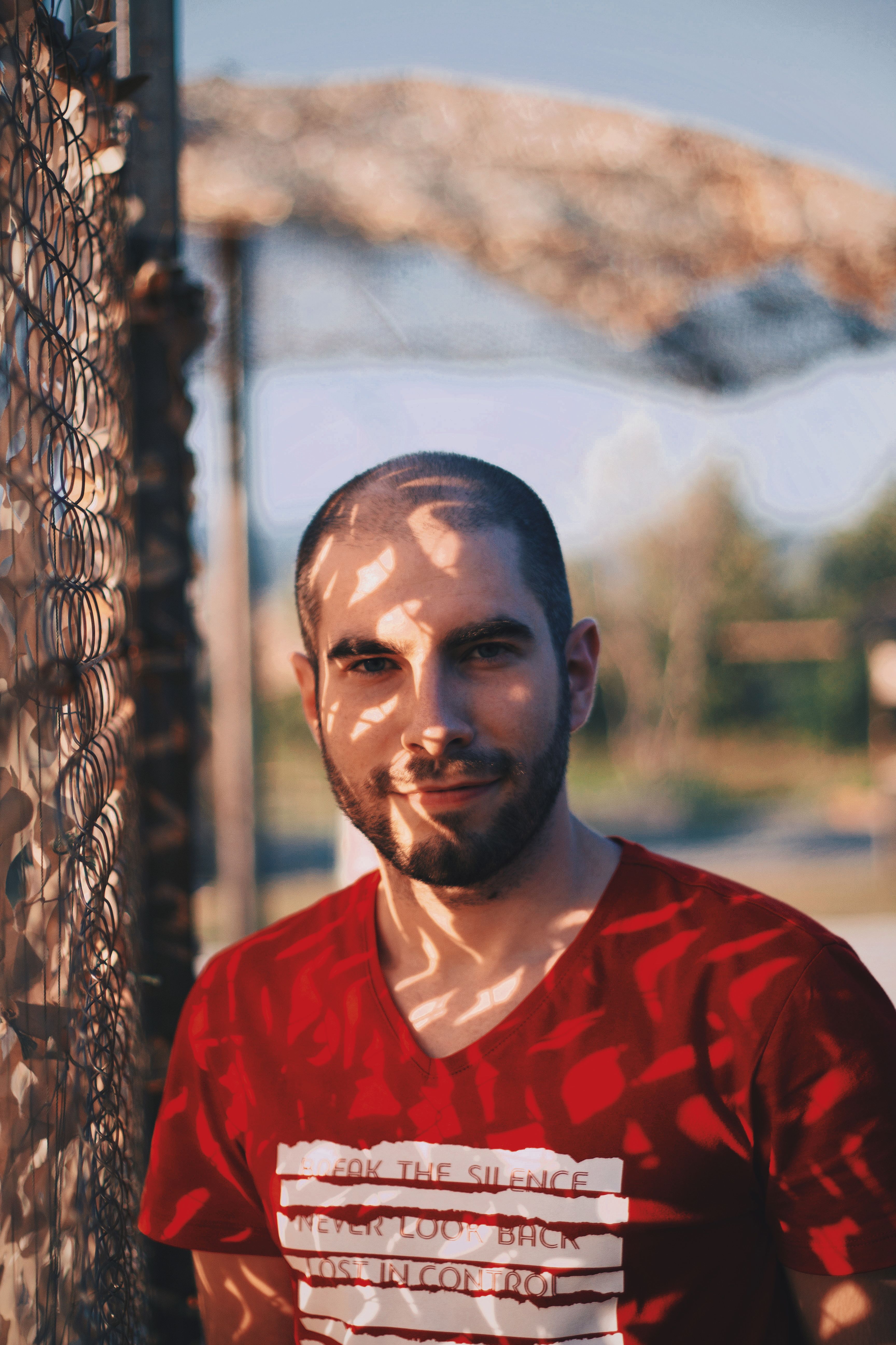 A bald man in a red shirt smiles by a chain-link fence outdoors.