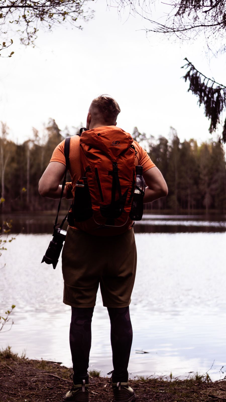 Man with orange backpack hiking along a tranquil forest lake trail, showcasing outdoor adventure. Stock Photo