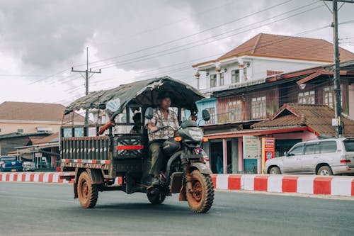 Ethnic male driver driving aged auto rickshaw on asphalt roadway near bright fence and buildings in town under cloudy sky