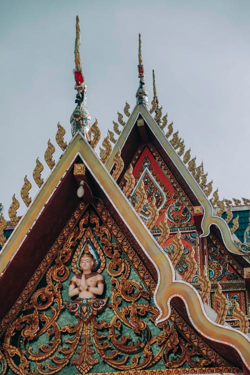 Low angle of aged ornamental church roof with small statue of Buddha and spiky peaks under sky