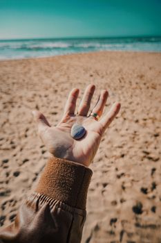 Hand holding a small stone on a sunny beach with gentle ocean waves.