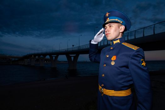 Serious man in navy uniform with shoulder strap and cap saluting while standing near bridge and river under cloudy sky in dusk and looking away