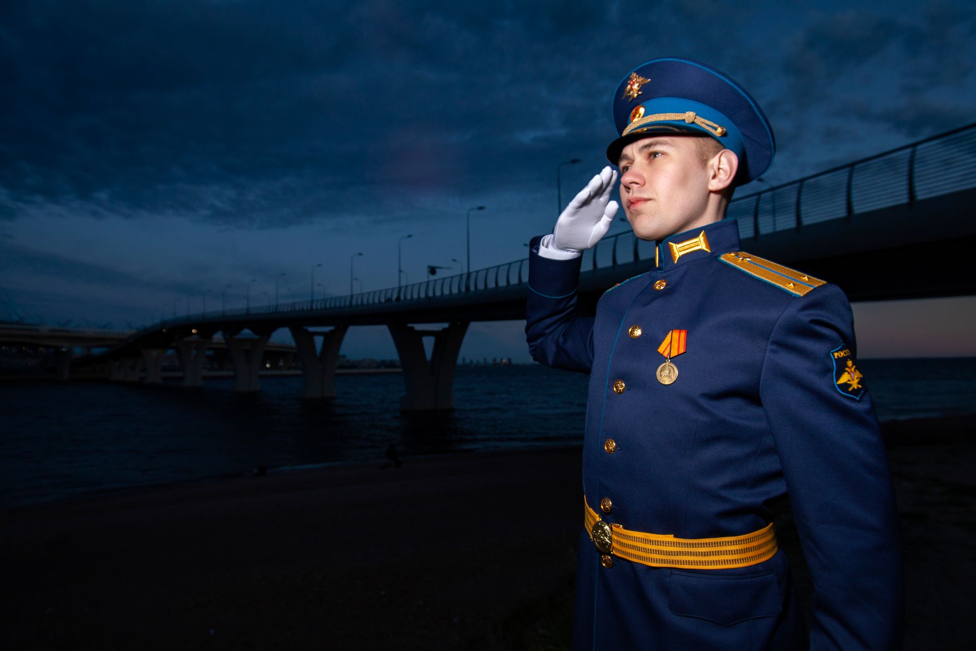 A young soldier in full uniform saluting by a bridge at dusk, symbolizing service and patriotism.