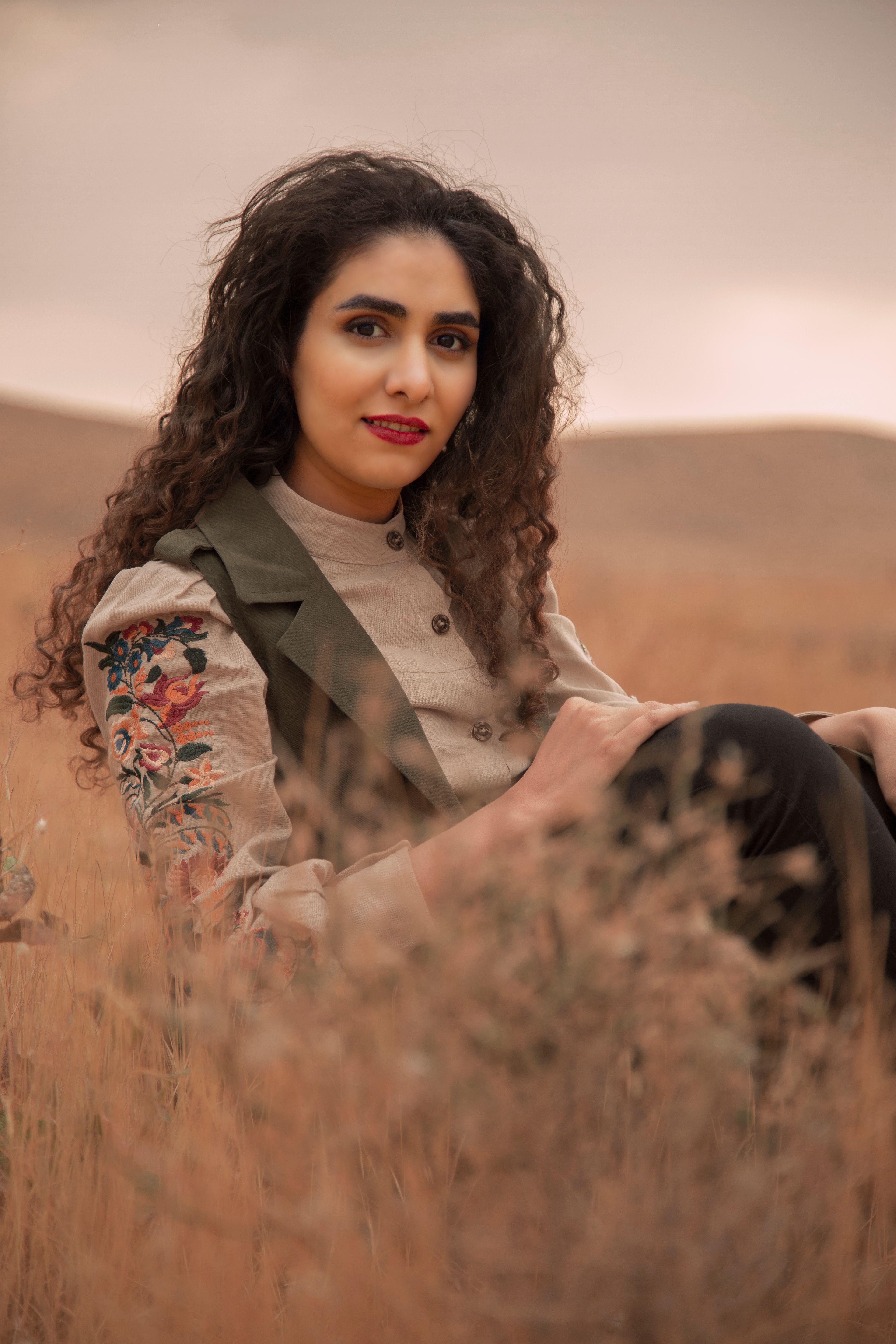 Portrait of a woman with curly hair and floral jacket posing in a warm, grassy field at sunset.