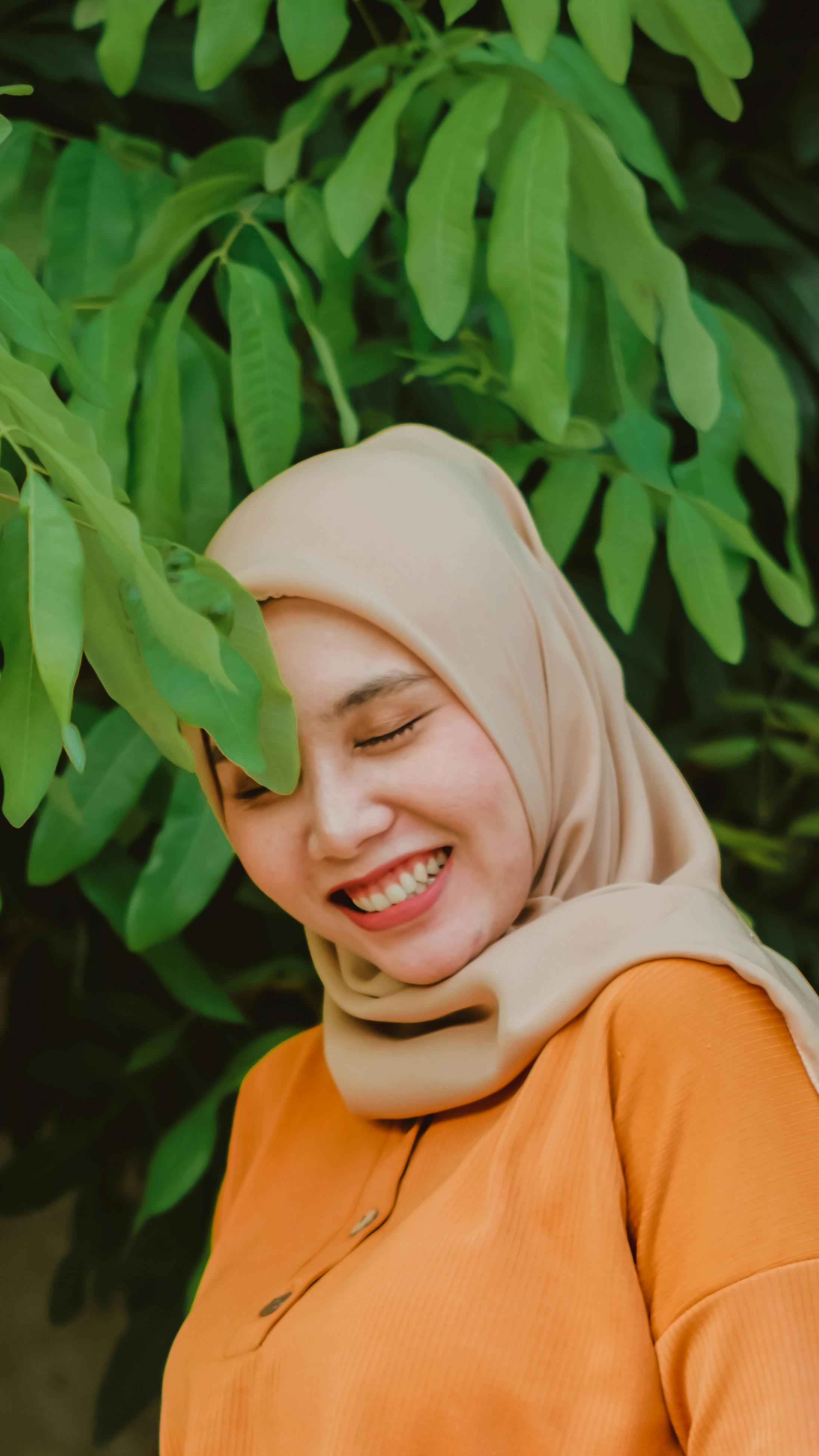 A joyful woman wearing a hijab stands in front of lush green leaves, smiling brightly.