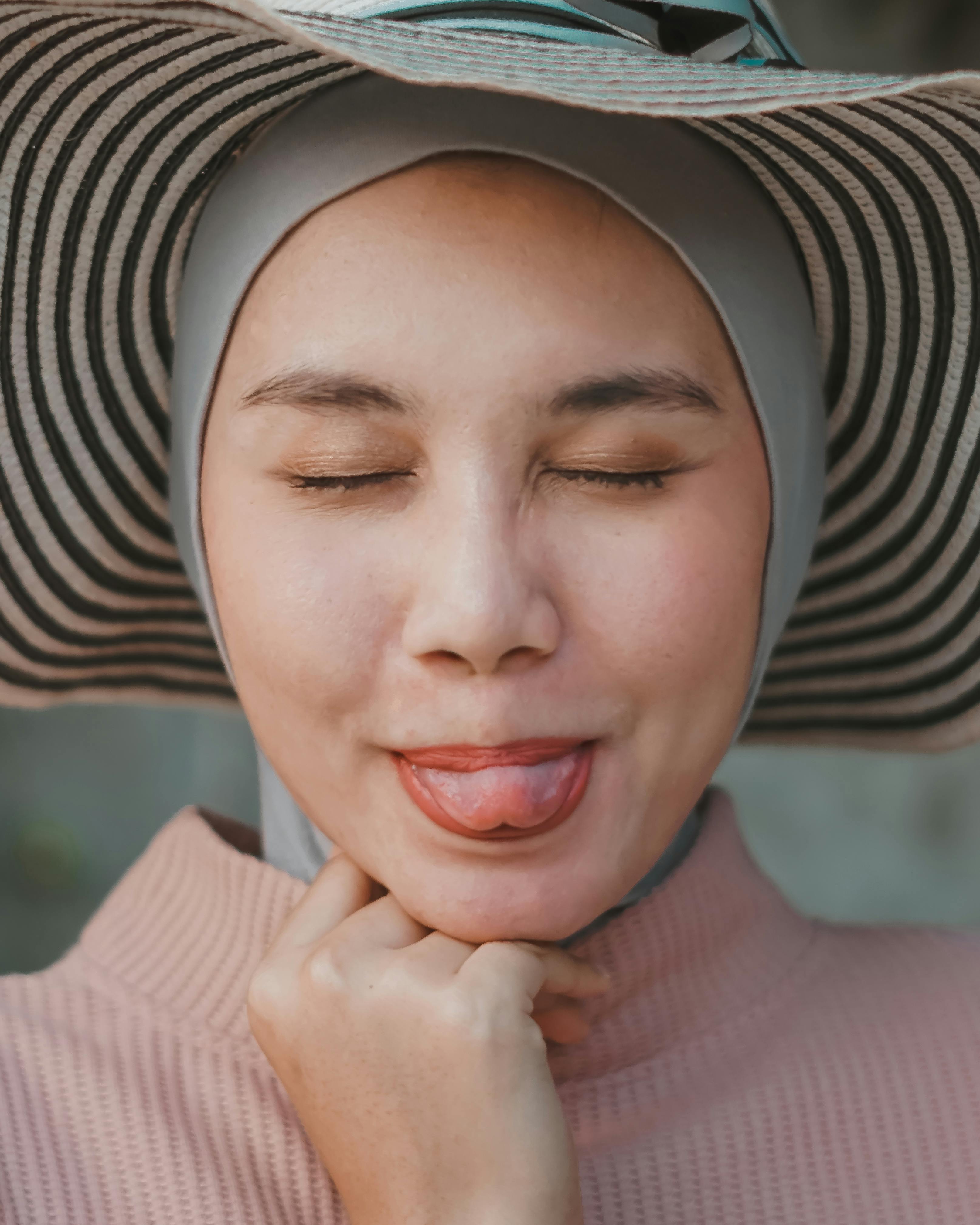 A woman in hijab and sun hat playfully sticking out her tongue with closed eyes.