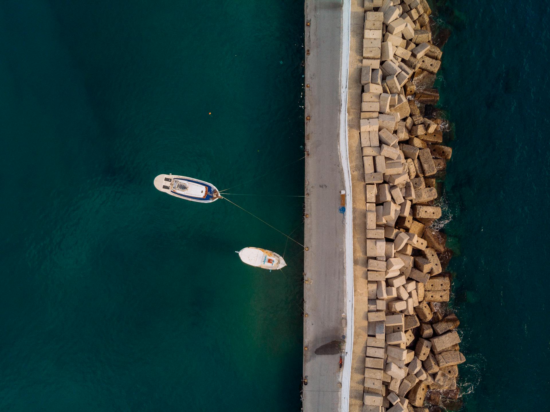 Drone shot of boats docked at a breakwater in a Greek harbor, showcasing the blue sea and concrete blocks.
