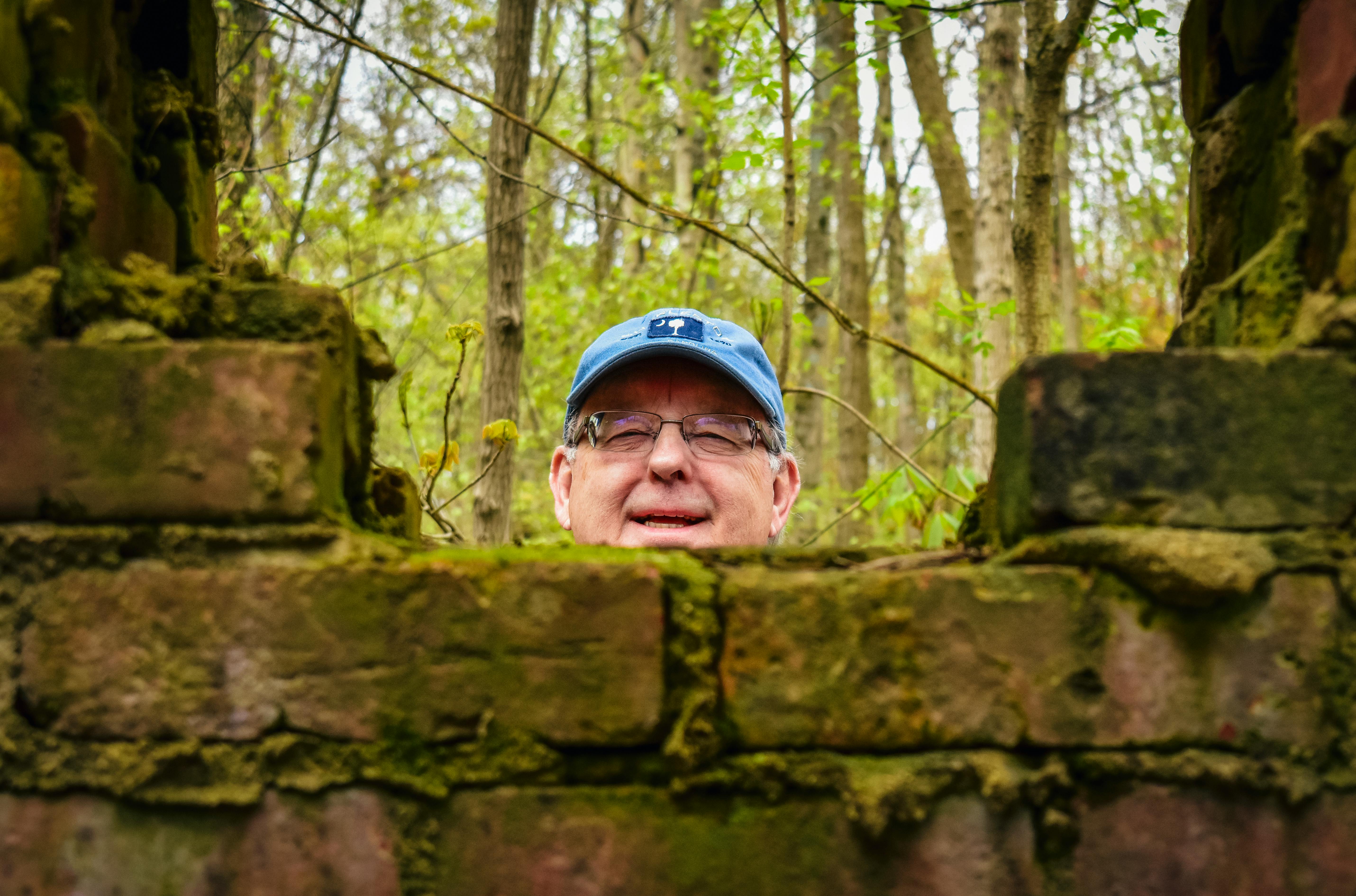 Smiling mature man peeking out from behind old brick fence · Free Stock  Photo