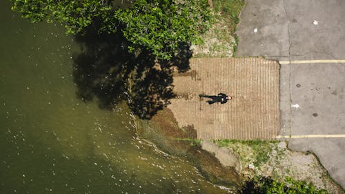 Free Overhead of anonymous male in black wear lying on old boardwalk between rippled river and cement pavement near trees Stock Photo