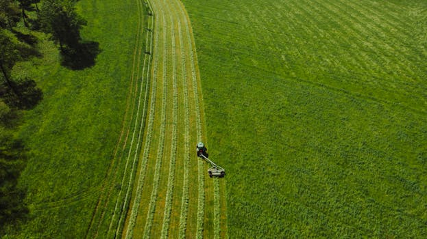 Drone captures tractor mowing field in lush green landscape from above.