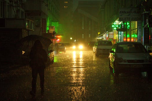 Silhouette of a person walking with an umbrella on a rainy city street at night, with car headlights.