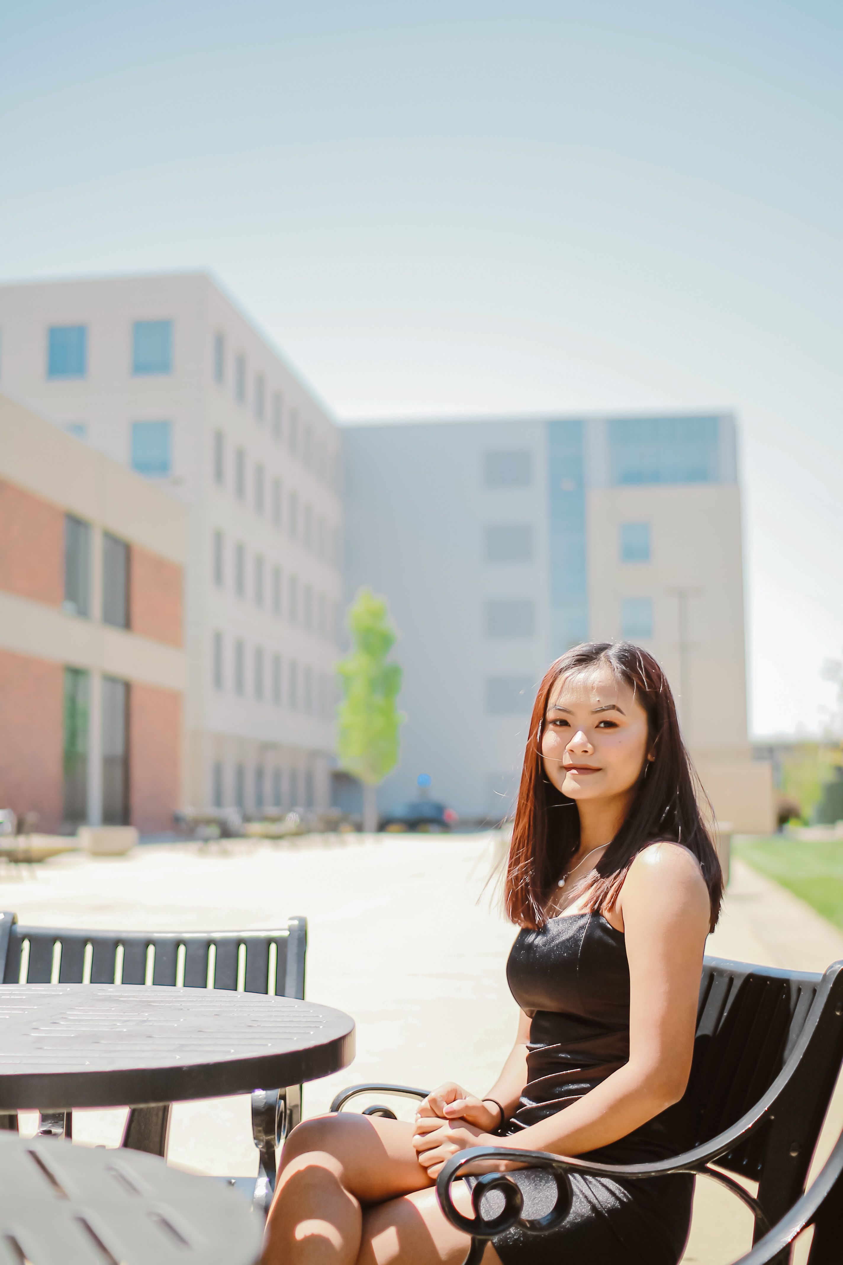 Young woman in black dress smiling while sitting outdoors on a sunny day at a university campus.