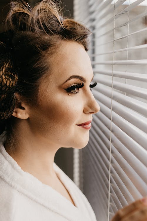 Side view of content female with bright makeup in white bathrobe looking at window pensively
