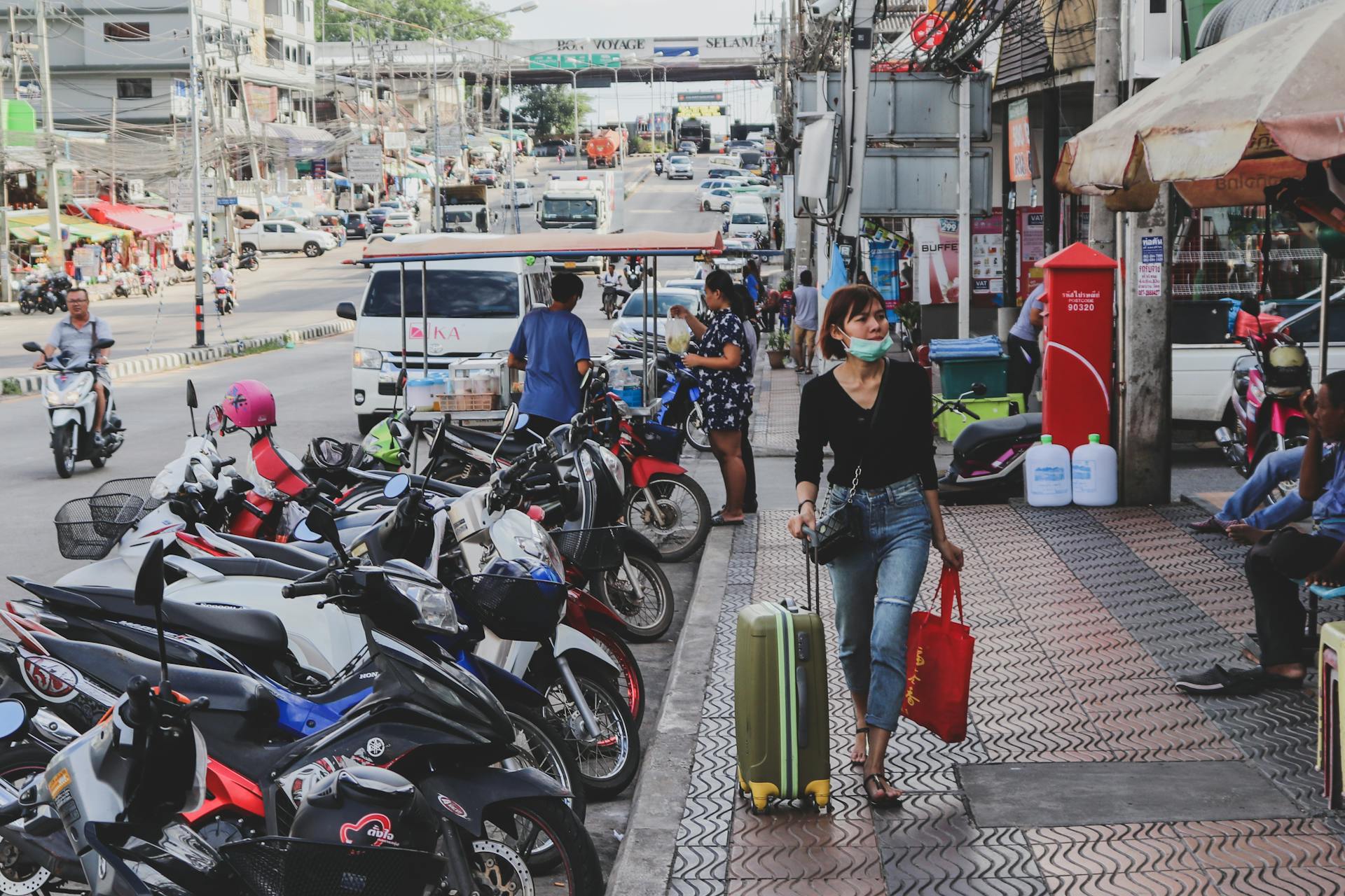 Woman carrying luggage and shopping bag on a busy city sidewalk. Motorbikes line the street.