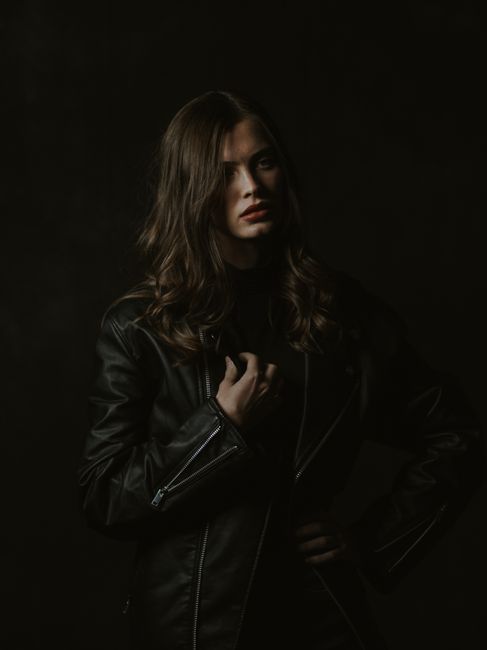 Serious slender brunette wearing trendy leather jacket standing on black background with hand on waist and looking away in dark studio