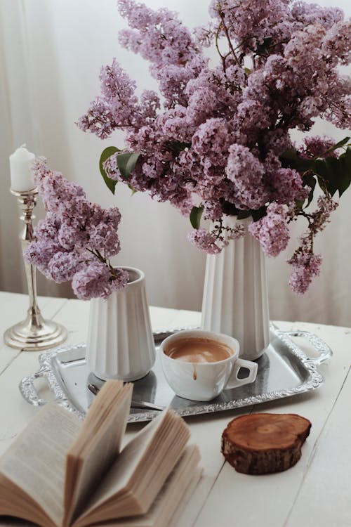 Free From above arrangement of fragrant lilac flowers in vase placed on tray with aromatic cappuccino served on table near opened book in light living room Stock Photo