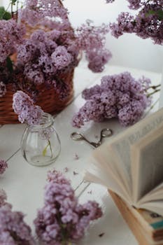 靠近堆放的书籍和罐子花瓶的桌子上，放着一个高角度排列的柳条篮，篮里盛开着丁香花