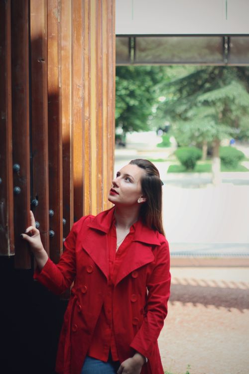 Free Caucasian woman in red coat gazes at architectural structure outdoors. Medium shot with trees in the background. Stock Photo