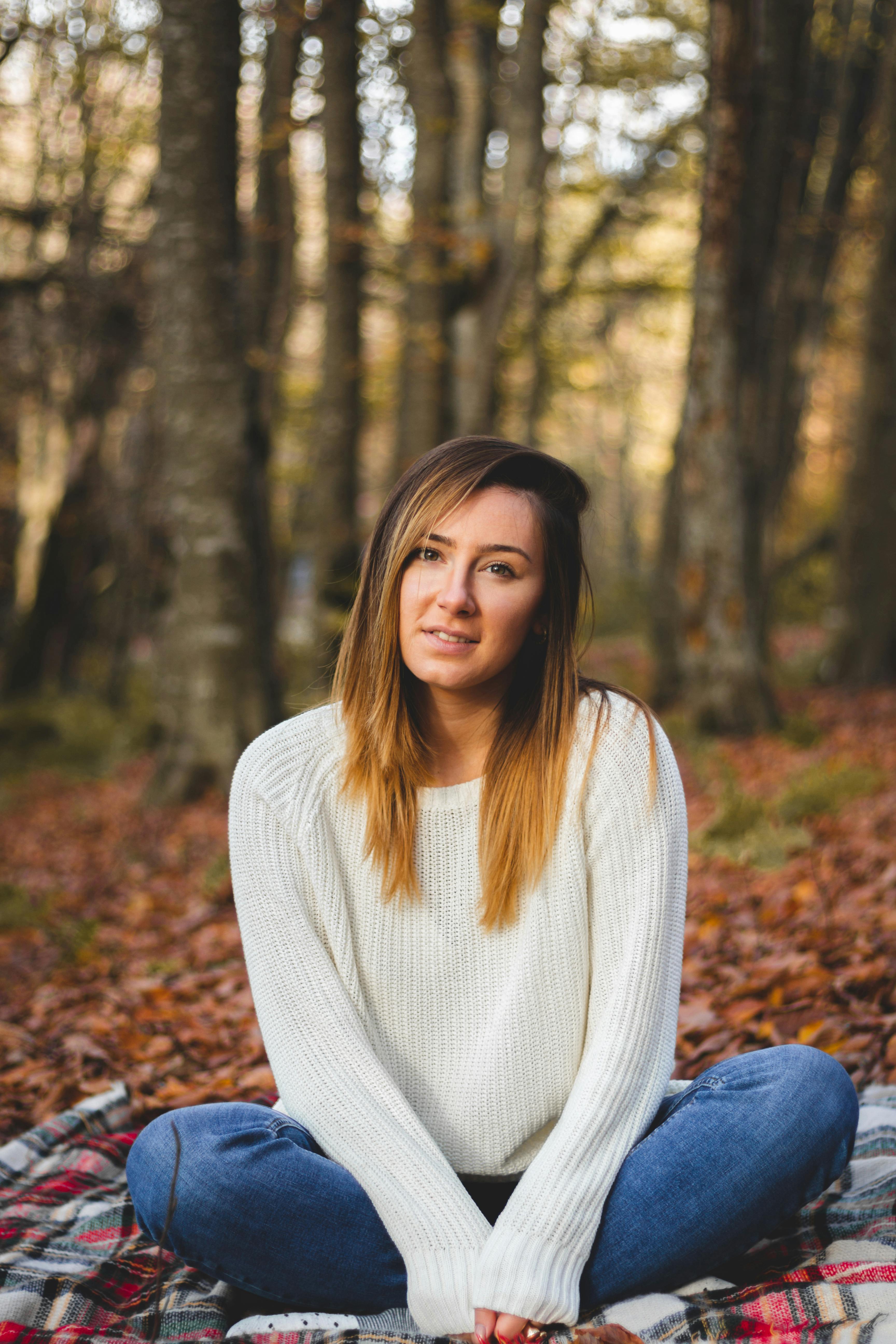 Free Caucasian woman in a white sweater sitting on a blanket in a serene autumn forest. Stock Photo