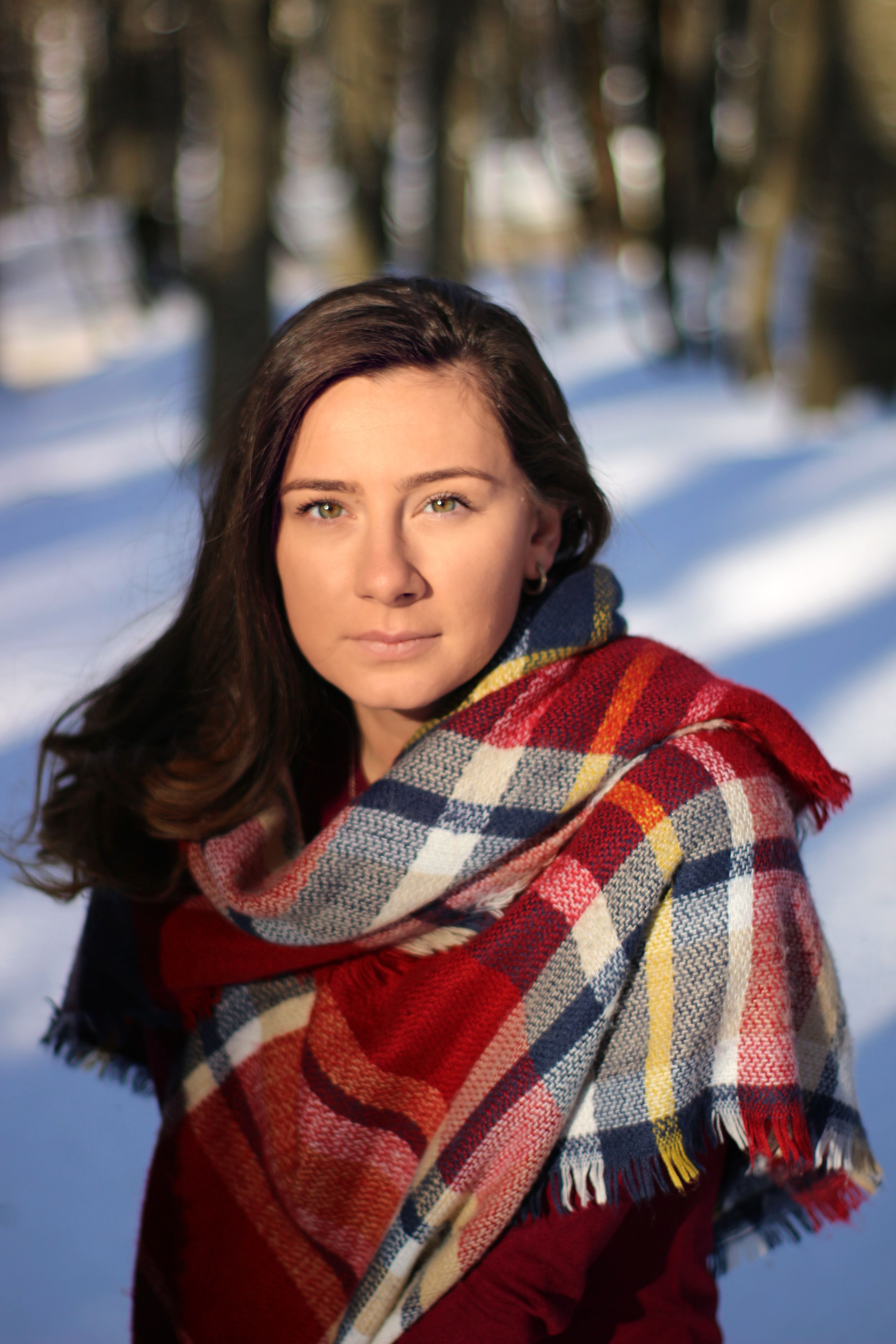A woman gazes into the distance while wrapped in a colorful plaid scarf, standing on a snowy day.