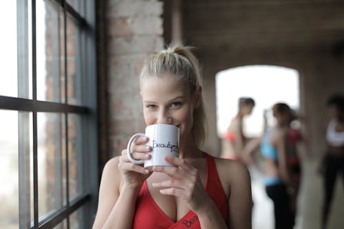 Smiling young blond female in sportswear looking at camera while standing by window in gym and drinking tea