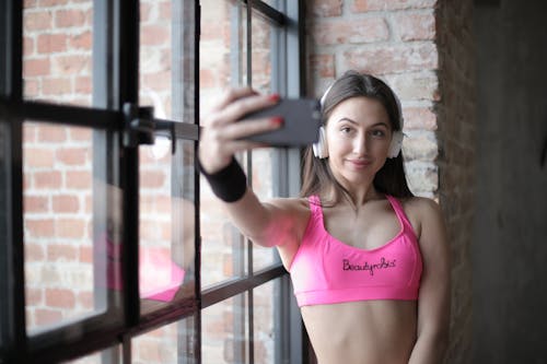 Positive slim female athlete wearing sports bra and headphones standing near window and smiling while looking at smartphone and taking selfie