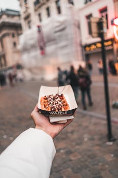 Hand holding a delicious Belgian waffle on a bustling Brussels street, highlighting local cuisine.