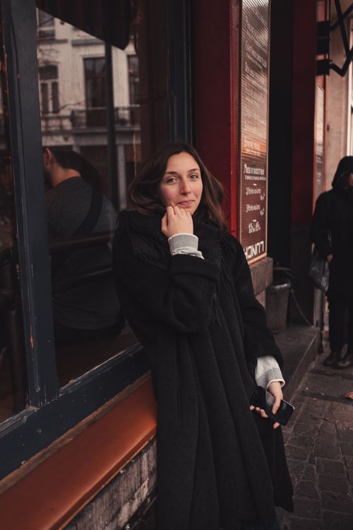 Free A woman in a black coat stands by a window in Brussels, reflecting city life. Stock Photo