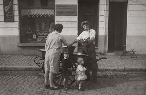 Grayscale Photo of A Mother And Her Children Buying Food From A Street Vendor