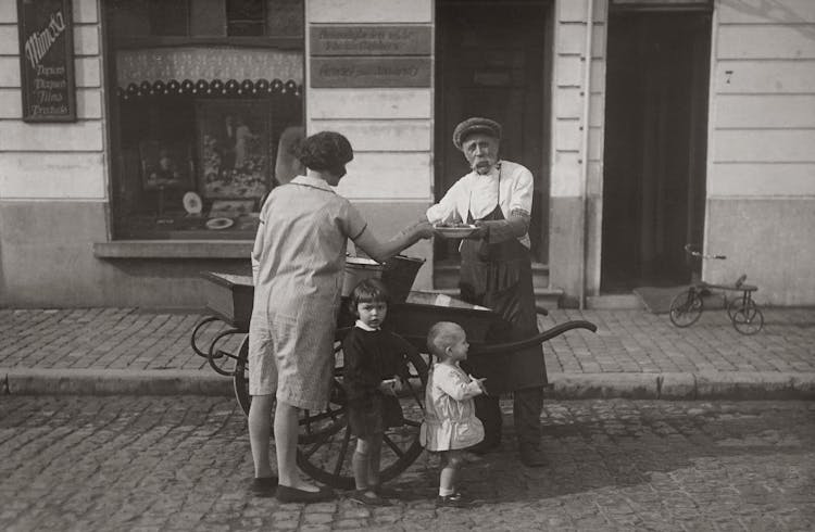 Grayscale Photo Of A Mother And Her Children Buying Food From A Street Vendor