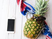 A pineapple with sunglasses and a colorful scarf next to a smartphone on a wooden table.