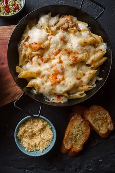 From above of yummy penne pasta in pan with sauce placed on table near grated Parmesan cheese and spicy toasted bread