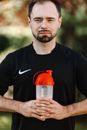 一名留着胡子、身穿黑衬衫的男子手持蛋白质瓶，站在户外，体现了健身与健康的理念