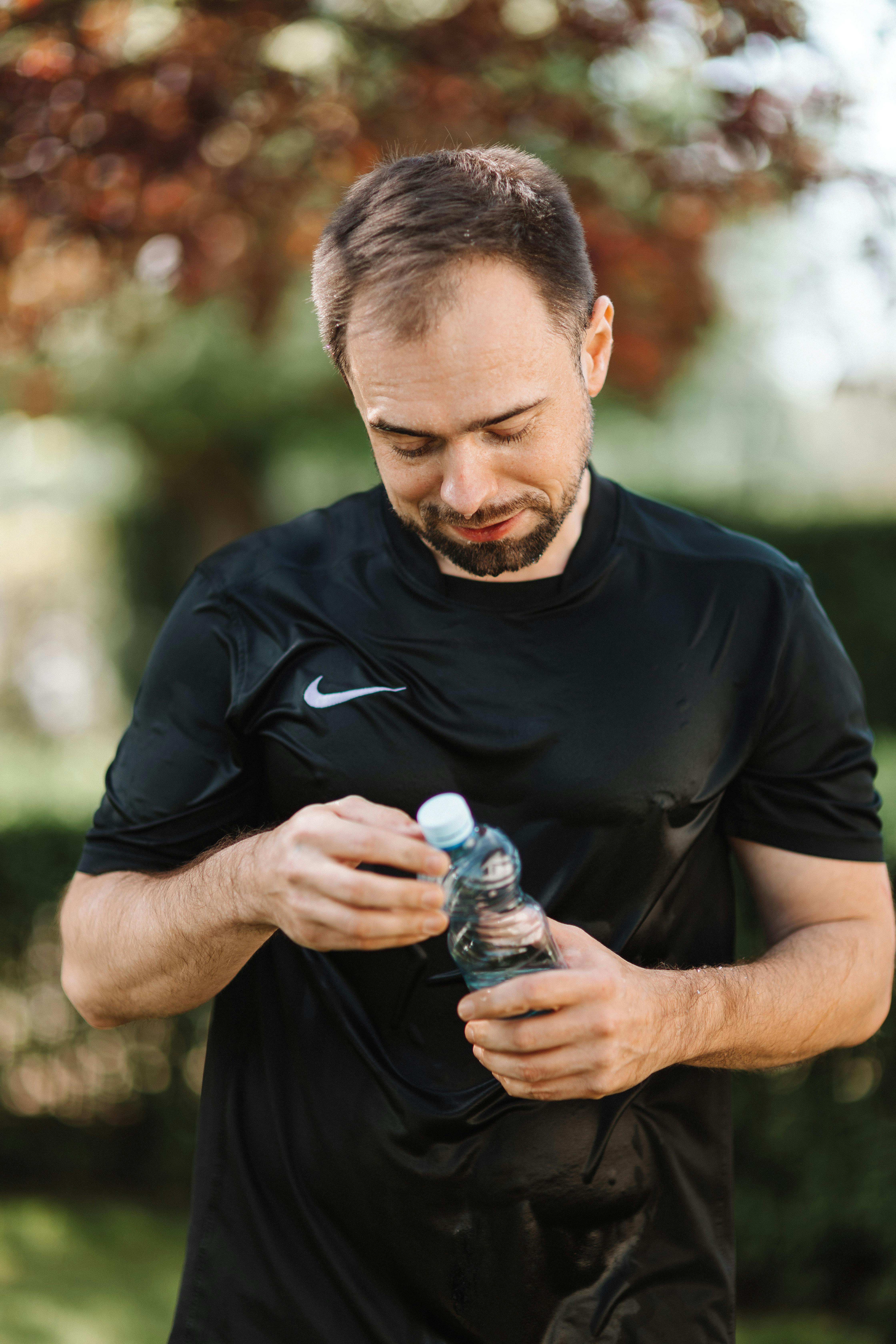 一名穿着黑色衬衫的男子在阳光明媚的户外打开塑料瓶，享受补水休息