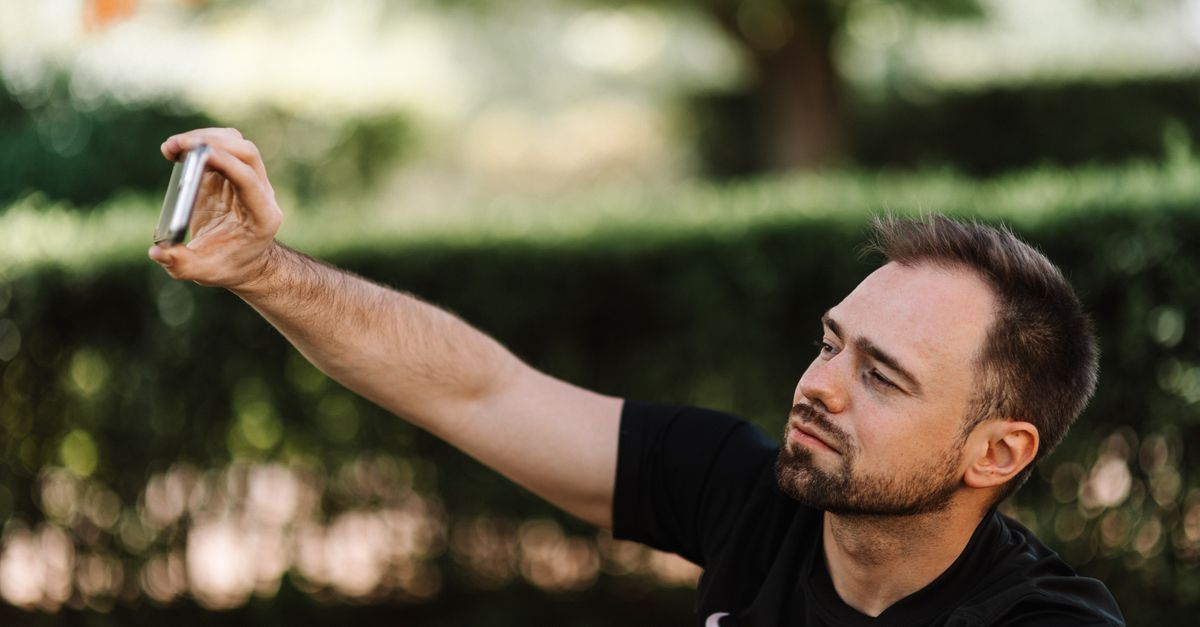 Young man taking a selfie with his smartphone in a sunny park setting.