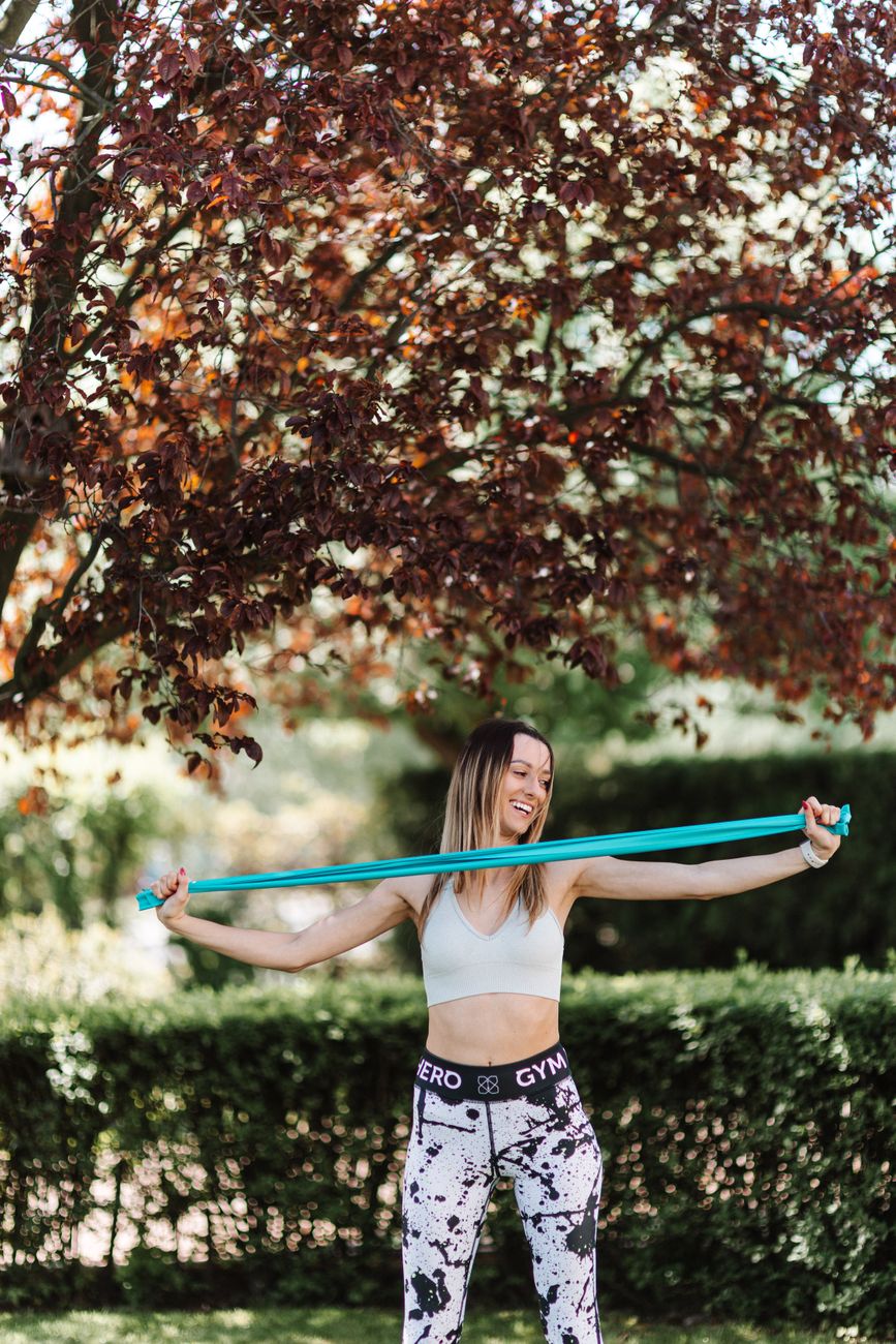 Smiling woman exercising with a resistance band outdoors under a tree.
