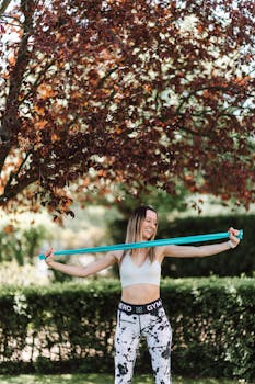 Smiling woman exercising with a resistance band outdoors under a tree.