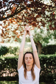 Woman practicing yoga outdoors with eyes closed, embracing tranquility under leafy trees.