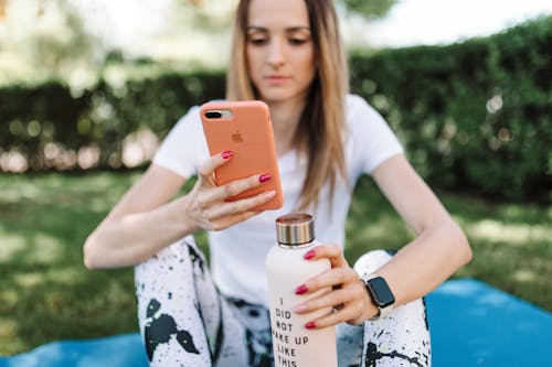 Shallow Focus Photo of a Woman Holding a Water Bottle while Using Her Iphone