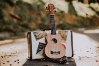 Wooden soprano ukulele placed on asphalt road with stylish suitcase on sunny autumn day