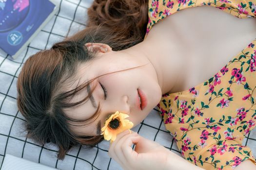 一位年轻的亚洲女性躺在毯子上，用新鲜的黄色花朵遮住眼睛，阳光明媚