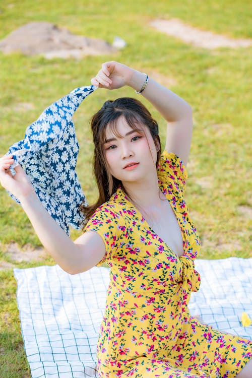 From above of positive young Asian female in stylish dress holding headscarf in raised hands while resting on green grass in park